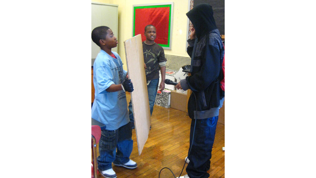 A youth looking at another youth holding a microphone looking at another youth holding a plank of wood
