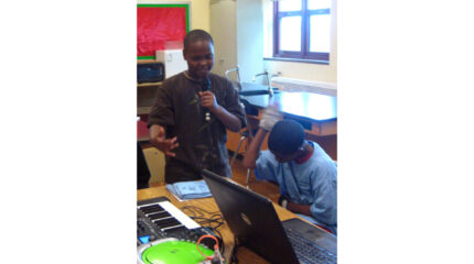 A youth sitting behind a laptop computer listening to another youth speaking into a held microphone in a classroom