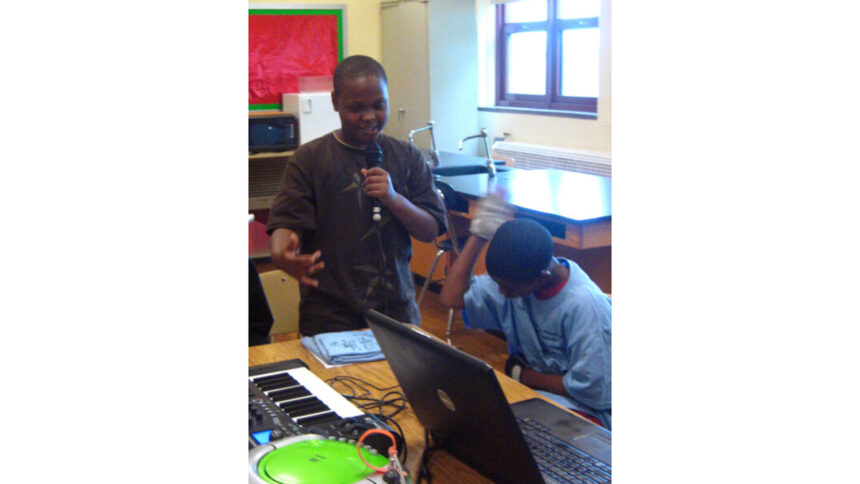 A youth sitting behind a laptop computer listening to another youth speaking into a held microphone in a classroom
