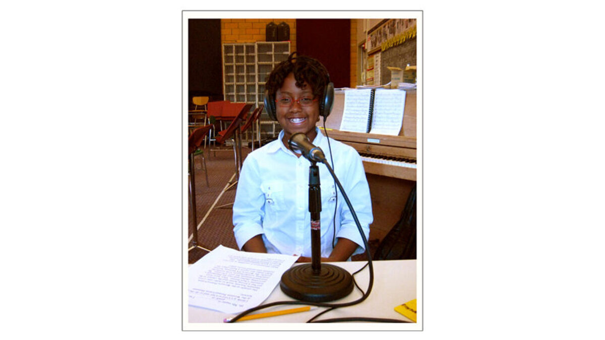 A youth wearing headphones smiling at the camera while sitting behind a microphone in a classroom