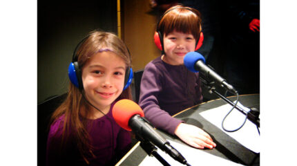 Two youth wearing headphones smiling and sitting behind microphones in a radio studio
