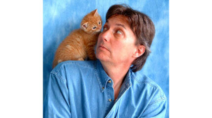 An adult looking at a cat sitting on their shoulder, both in front of a blue background
