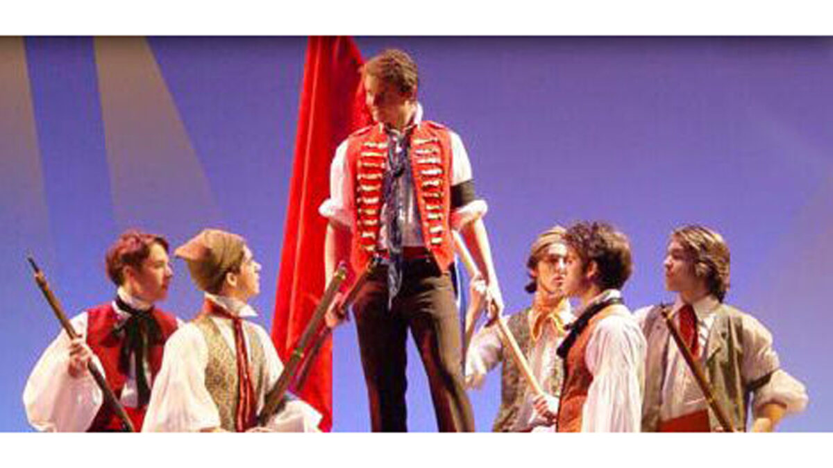 Six individuals dressed in late 1700's garb, one holding a large red flag, the rest holding muskets