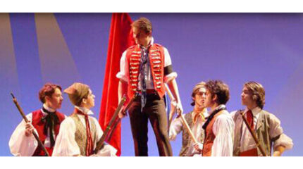 Six individuals dressed in late 1700's garb, one holding a large red flag, the rest holding muskets