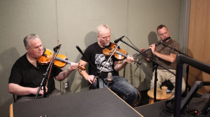 Two adults playing violins and an adult playing a tin whistle in a radio studio