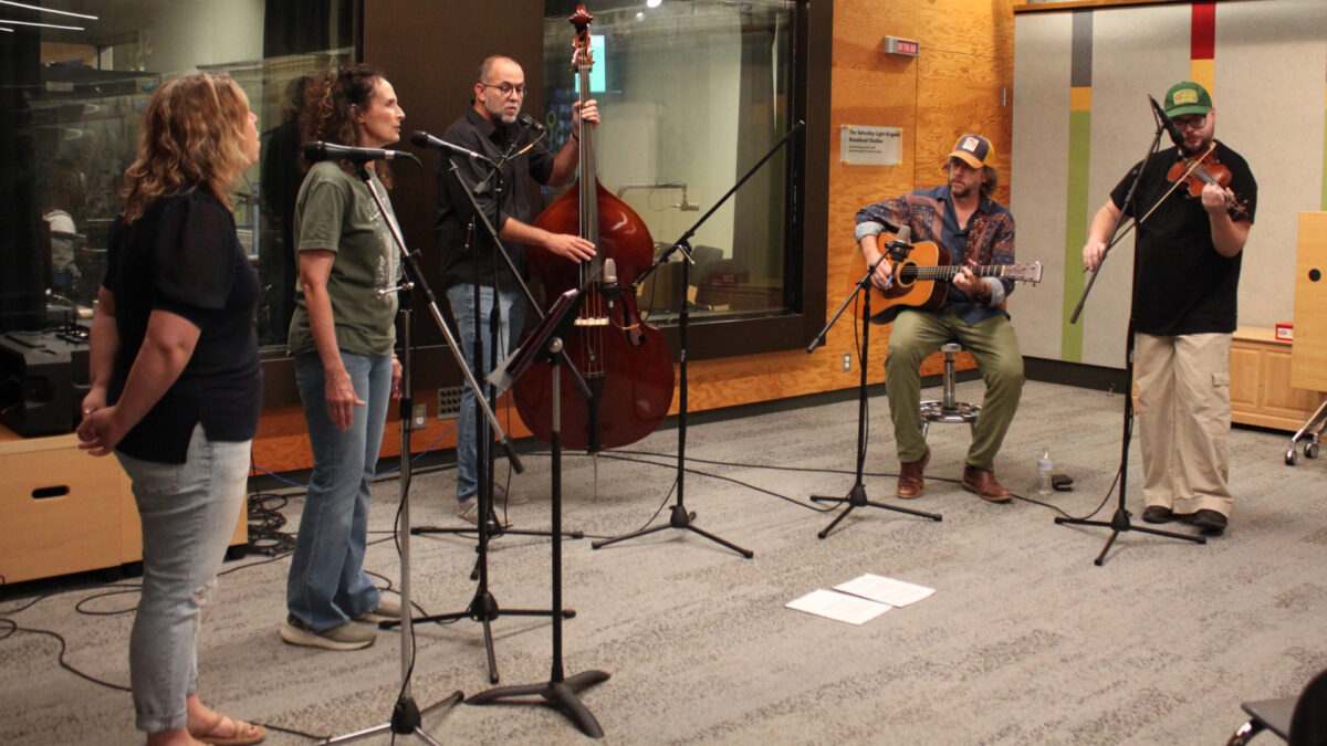 City-Holler-Band-4 Two adults singing into microphones, an adult playing upright bass and singing into a microphone, an adult playing acoustic guitar, and an adult playing fiddle