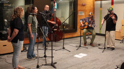 Two adults singing into microphones, an adult playing upright bass and singing into a microphone, an adult playing acoustic guitar, and an adult playing fiddle