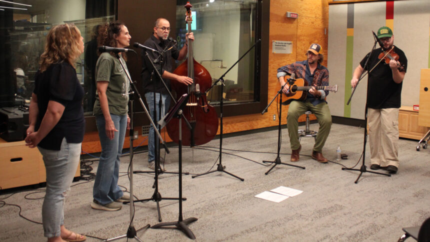 Two adults singing into microphones, an adult playing upright bass and singing into a microphone, an adult playing acoustic guitar, and an adult playing fiddle
