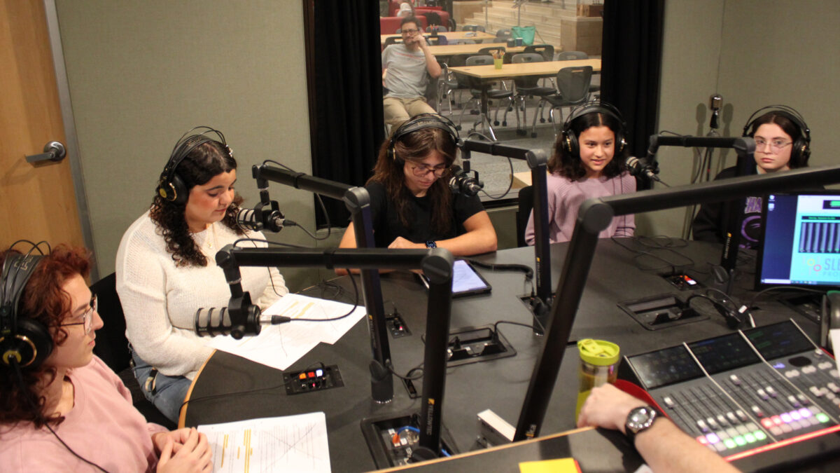 Five teens wearing headphones sitting behind microphones being interviewed in a radio studio