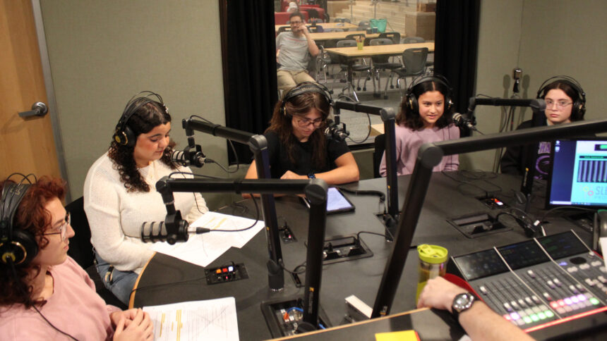 Five teens wearing headphones sitting behind microphones being interviewed in a radio studio