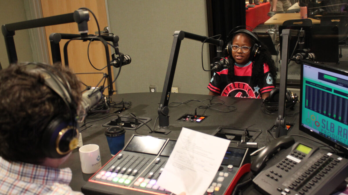 A youth wearing headphones speaking into a microphone being interviewed by an adult wearing headphones sitting behind an audio mixer in a radio studio