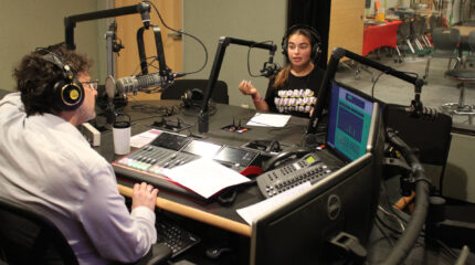 A teen wearing headphones speaking into a microphone being interviewed by an adult wearing headphones and sitting behind a microphone and an audio mixer in a radio studio