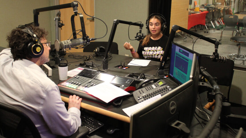 A teen wearing headphones speaking into a microphone being interviewed by an adult wearing headphones and sitting behind a microphone and an audio mixer in a radio studio