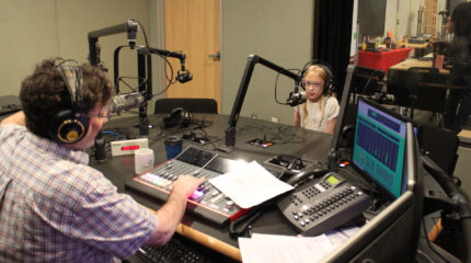 A youth wearing headphones sitting behind a microphone being interviewed by an adult wearing headphones sitting behind a microphone and an audio mixer in a radio studio