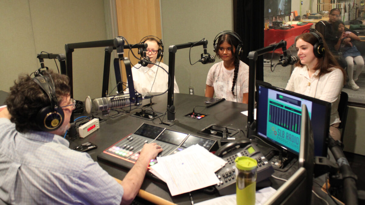 Three teens wearing headphones sitting behind microphones being interviewed by an adult wearing headphones speaking into a microphone in a radio studio