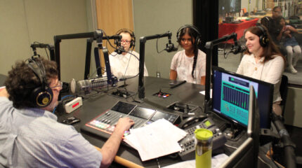Three teens wearing headphones sitting behind microphones being interviewed by an adult wearing headphones speaking into a microphone in a radio studio