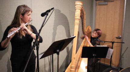 A teen playing flute and a teen playing harp, both looking at sheet music on music stands in a radio studio