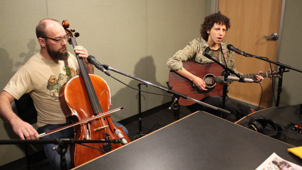 An adult playing cello and an adult playing acoustic guitar and singing into a microphone in a radio studio