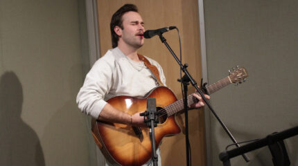 An adult playing acoustic guitar and singing into a microphone in a radio studio