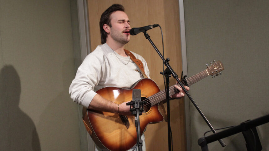 An adult playing acoustic guitar and singing into a microphone in a radio studio