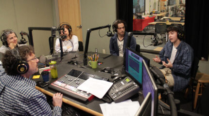 Three adults and a teen wearing headphones speaking into microphones being interviewed by an adult wearing headphones sitting behind a microphone and an audio mixer in a radio studio