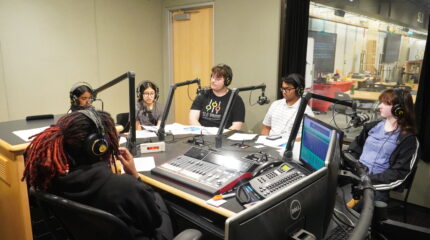 Five teens wearing headphones speaking into microphones as an adult wearing headphones sitting behind an audio mixer listens in a radio studio