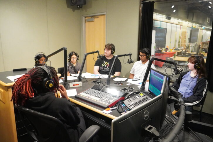 Five teens wearing headphones speaking into microphones as an adult wearing headphones sitting behind an audio mixer listens in a radio studio
