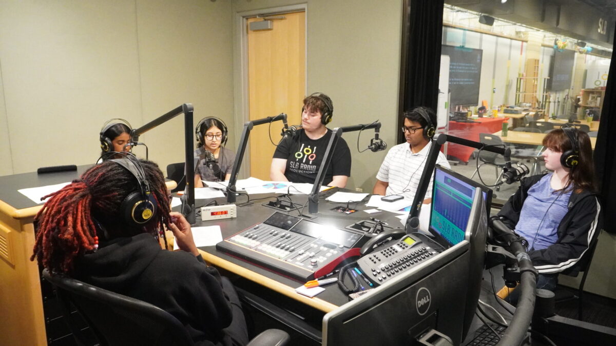 Streaming-Labor-2026_4 Five teens wearing headphones speaking into microphones as an adult wearing headphones sitting behind an audio mixer listens in a radio studio