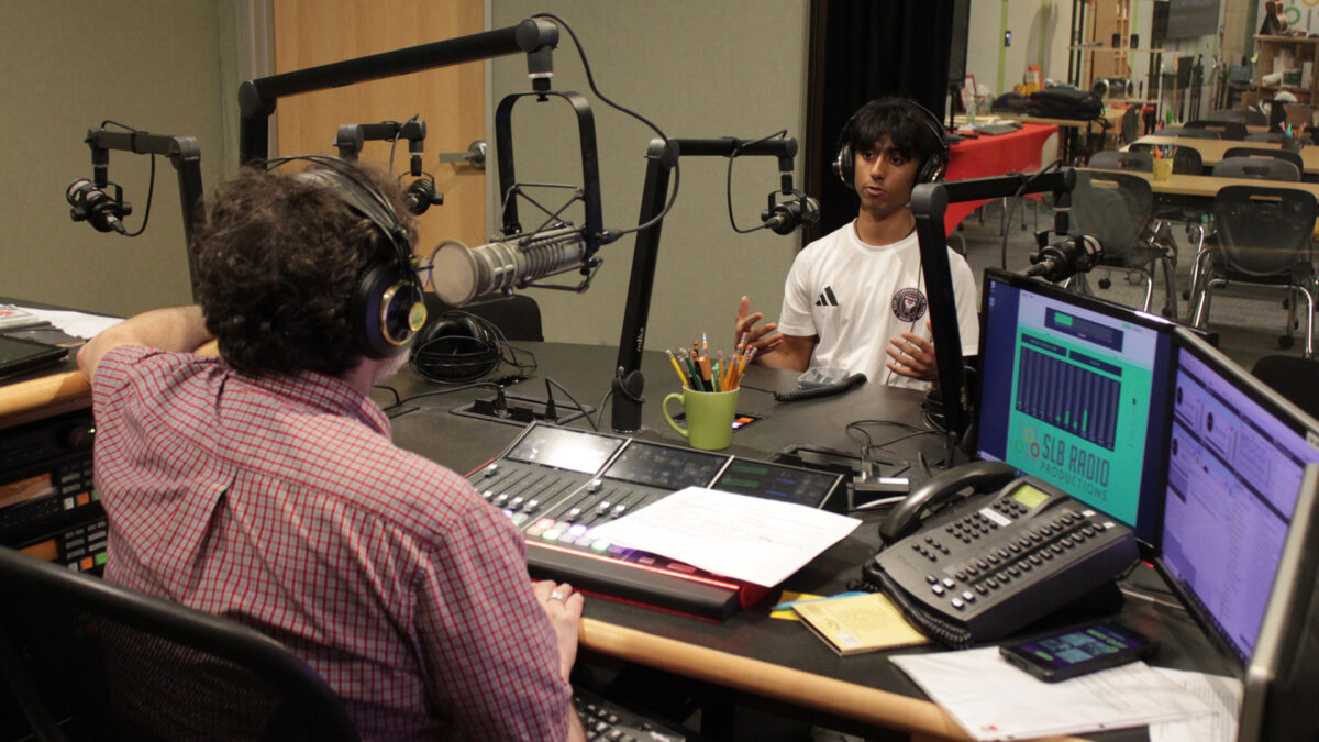 A teen wearing headphones speaking into a microphone being interviewed by an adult wearing headphones and sitting behind a microphone and an audio mixer in a radio studio