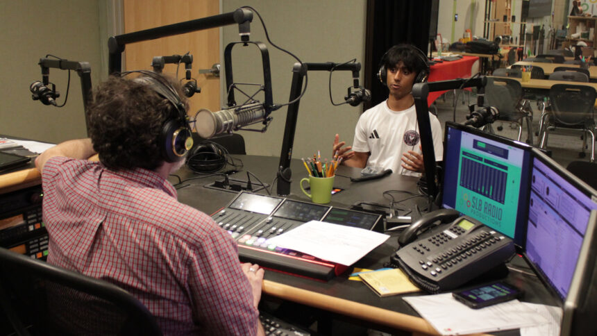 A teen wearing headphones speaking into a microphone being interviewed by an adult wearing headphones and sitting behind a microphone and an audio mixer in a radio studio