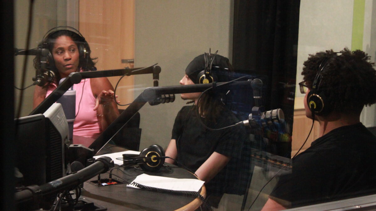 An adult and two teens wearing headphones and speaking into microphones in a radio studio
