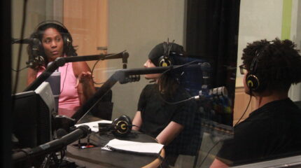 An adult and two teens wearing headphones and speaking into microphones in a radio studio