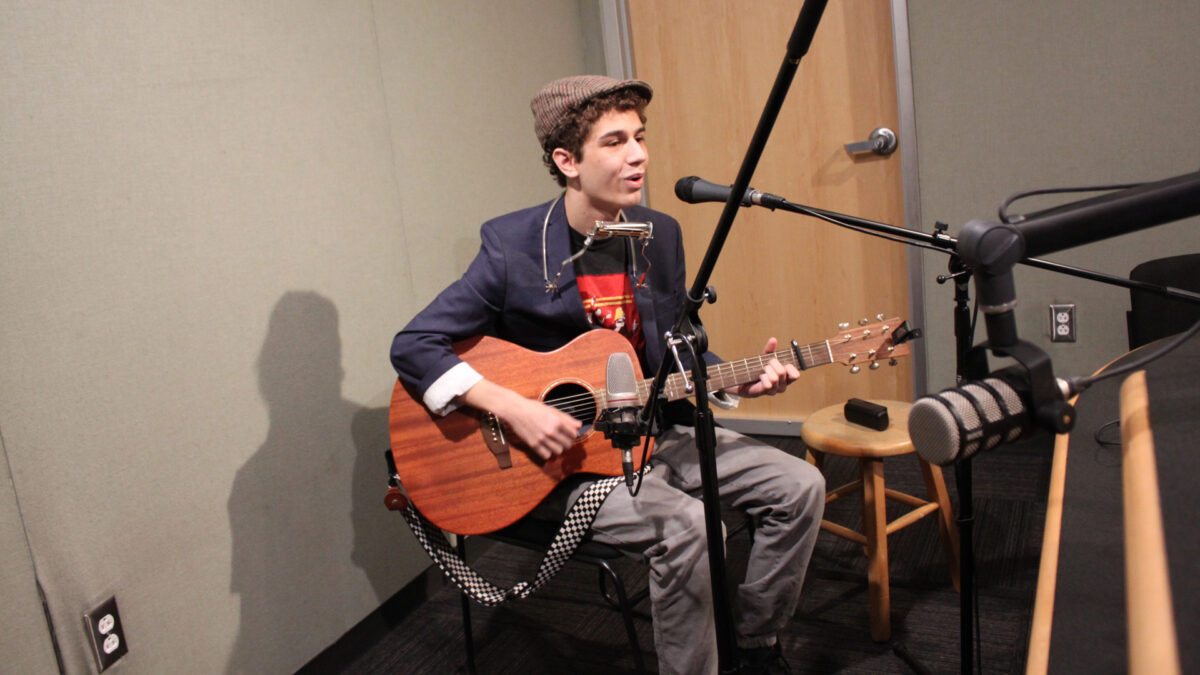 A teen playing acoustic guitar and singing into a microphone in a radio studio