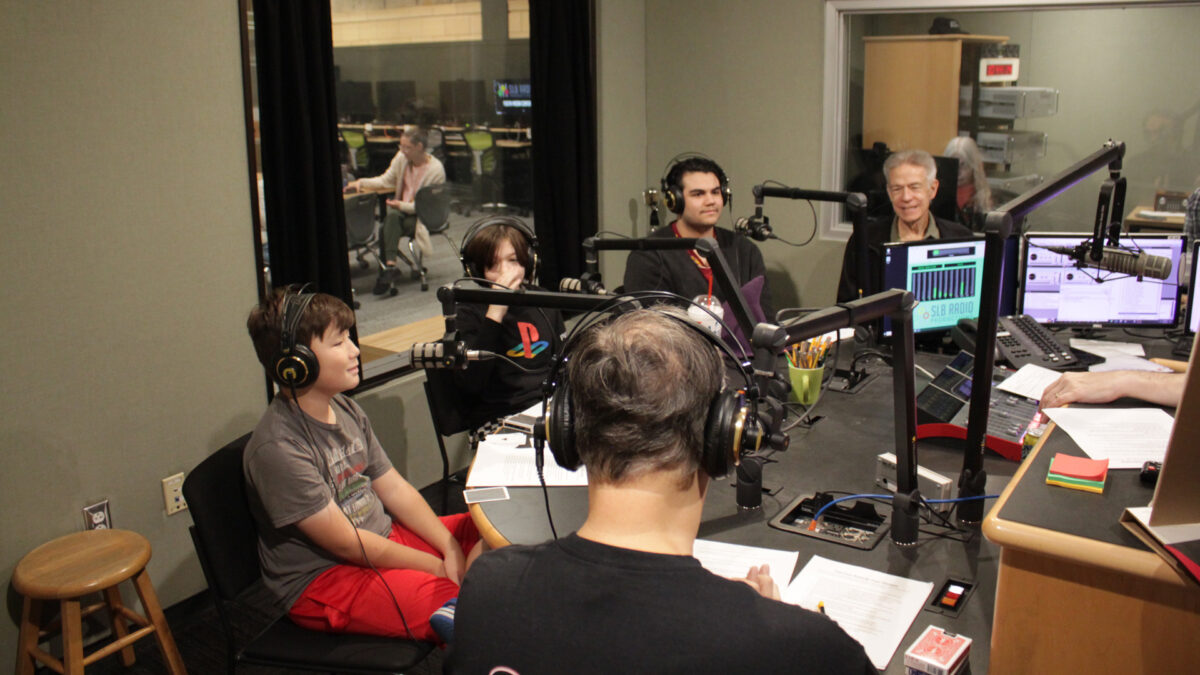 Three youth and two adults wearing headphones sitting behind microphones being interviewed in a radio studio