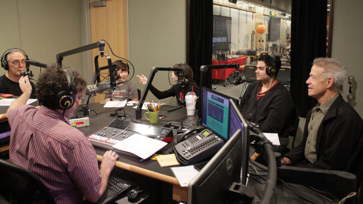 Three youth and two adults wearing headphones sitting behind microphones being interviewed by an adult wearing headphones sitting behind a microphone and an audio mixer in a radio studio