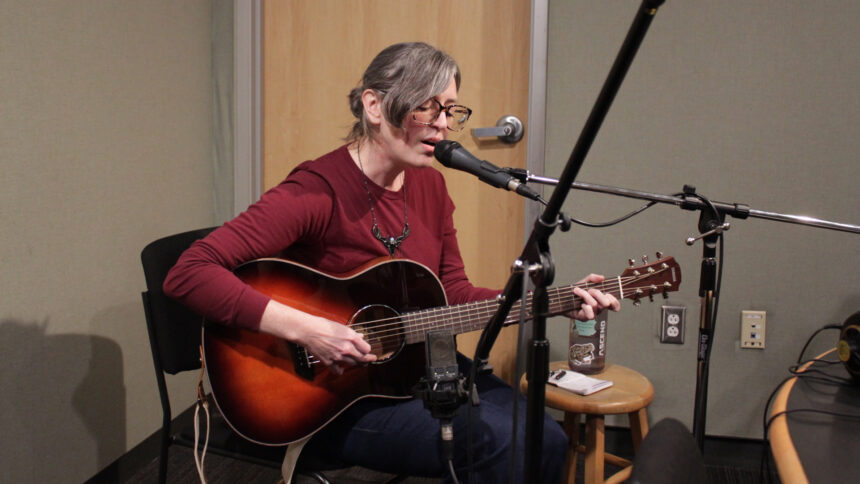 An adult playing acoustic guitar and singing into a microphone in a radio studio