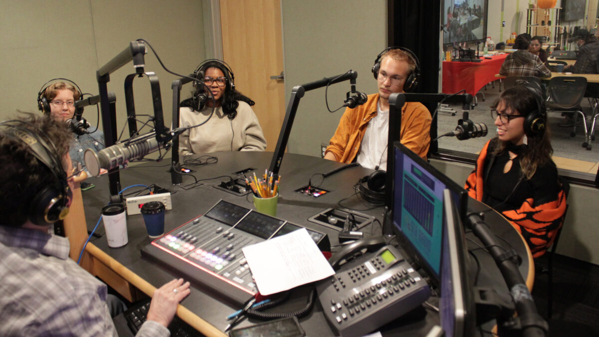 Three teens and one adult wearing headphones speaking into microphones being interviewed by an adult wearing headphones sitting behind a microphone and an audio mixer in a radio studio