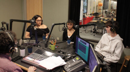 Three teens wearing headphones speaking into microphones being interviewed by an adult wearing headphones sitting behind a microphone and an audio mixer in a radio studio