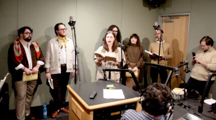 Six adults standing behind microphones, one singing, as another adult films on a smartphone and another adult wearing headphones listens in a radio studio