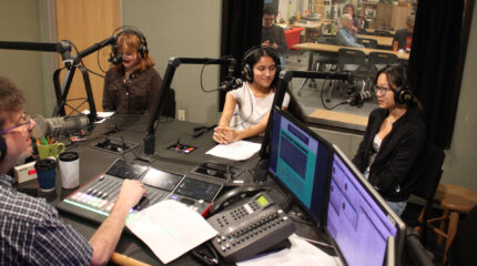 Three teens wearing headphones speaking into microphones being interviewed by an adult sitting behind a microphone and an audio mixer in a radio studio