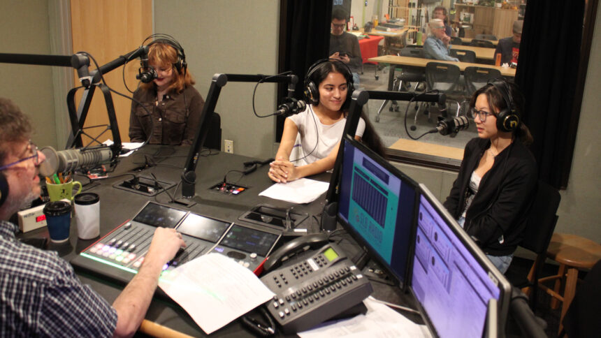 Three teens wearing headphones speaking into microphones being interviewed by an adult sitting behind a microphone and an audio mixer in a radio studio