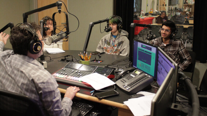 Three teens wearing headphones speaking into microphones being interviewed by an adult wearing headphones sitting behind a microphone and an audio mixer in a radio studio