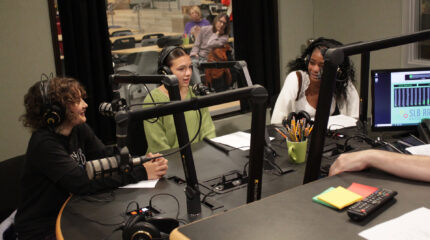 Three teens wearing headphones speaking into microphones in a radio studio