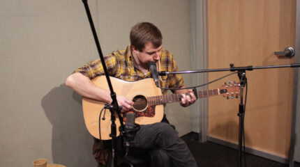 An adult playing acoustic guitar and singing into a microphone in a radio studio